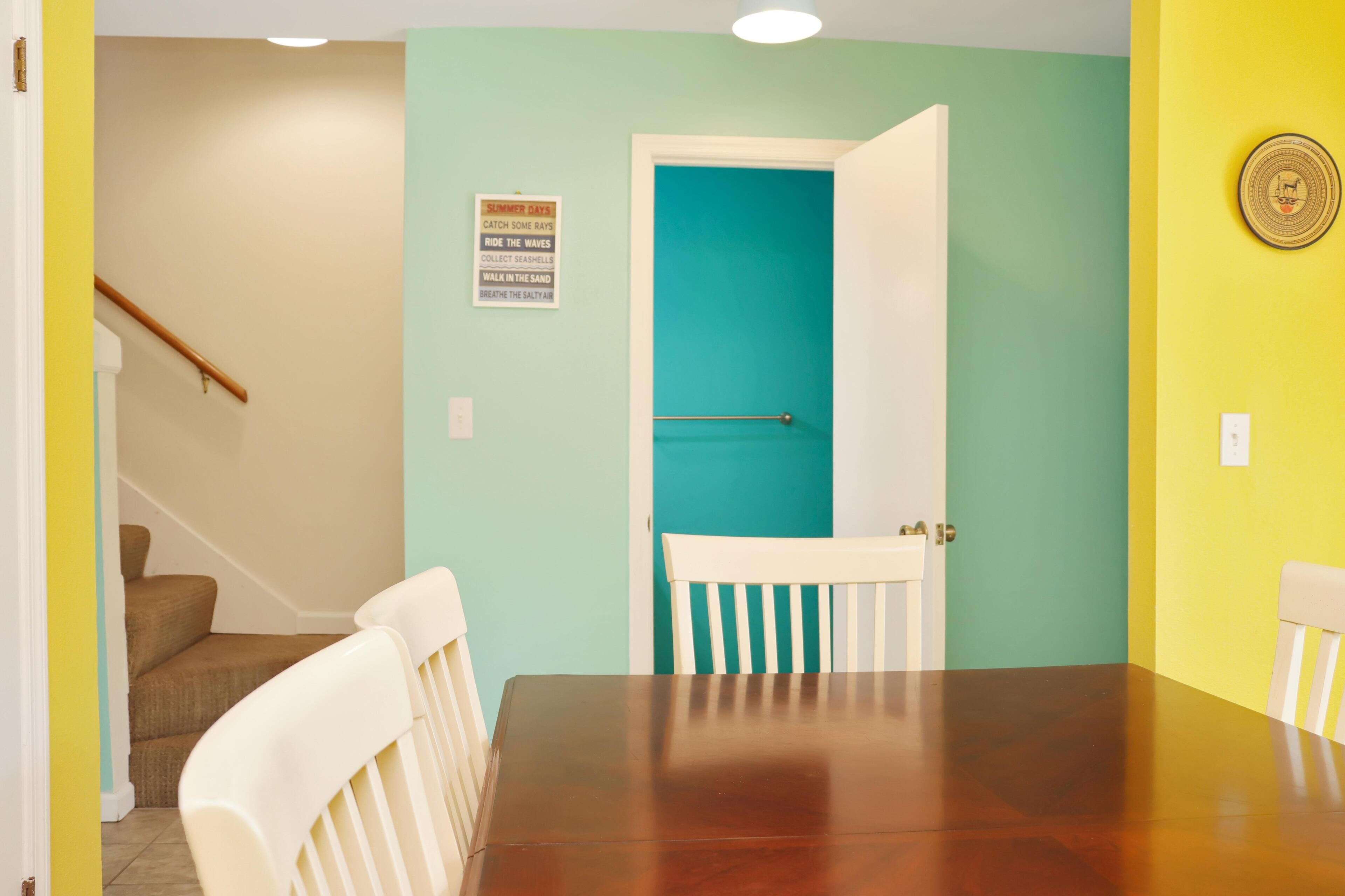 A dining area with a wooden table and white chairs, a teal door, and a staircase in the background.