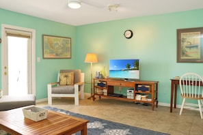 A living room with a wooden coffee table, a white armchair, a desk with a lamp, a television, and a framed map on the wall. - Sea Cliff B-2 (Port St. Joe)