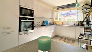 A modern kitchen with white cabinets, a double oven, and a window with a view of a house. - 5 minutes from the beach, quiet renovated house with wifi, air conditioning and jaccuzi (CANET EN ROUSSILLON)