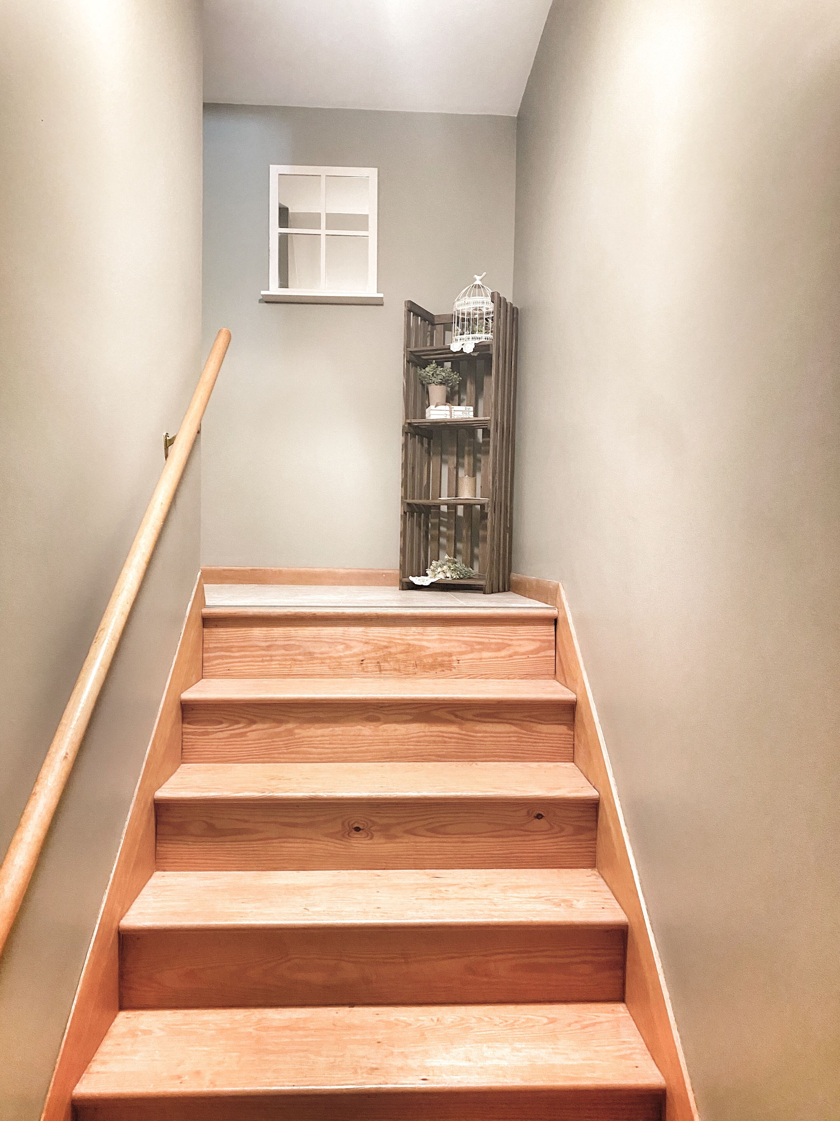 Un escalier en bois avec une rampe, une petite fenêtre et une bibliothèque en bois ornée d’objets décoratifs.