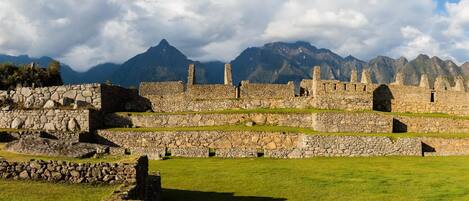 A historical stone structure with multiple terraces and ruins, surrounded by mountains and greenery.