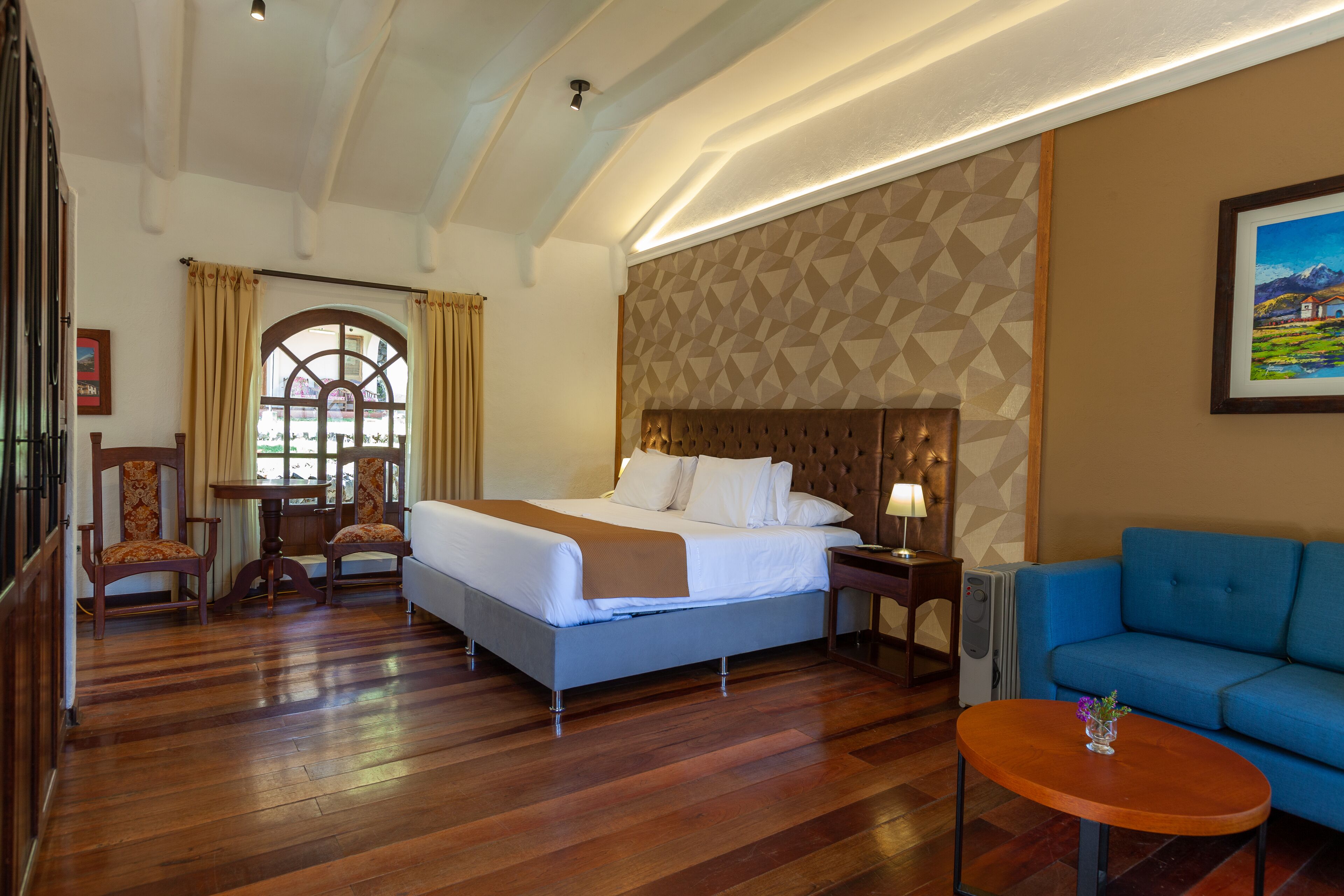 A hotel room with a large bed, a blue sofa, a wooden coffee table, and a small table with chairs.