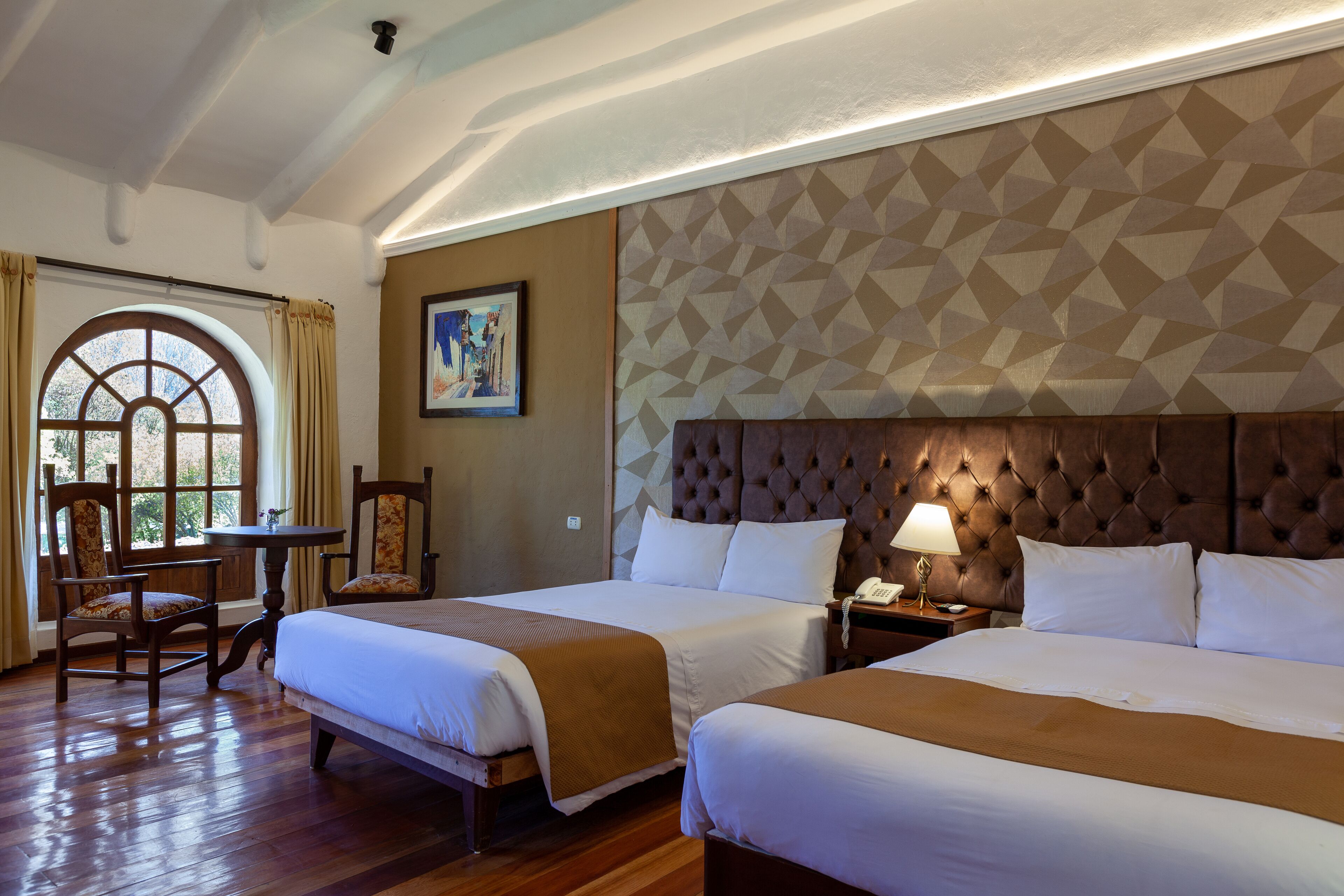 A hotel room with two beds, a large window with curtains, a round table, and a painting on the wall.
