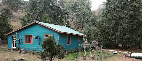 A blue house with a red door and windows, surrounded by trees and a swing set.