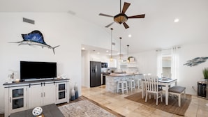 A modern kitchen with white cabinets, a central island with bar stools, a ceiling fan, and a large flat-screen TV.
