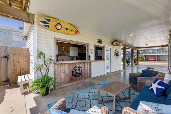 A covered outdoor seating area with a wooden table, wicker furniture, and a surfboard decoration.
