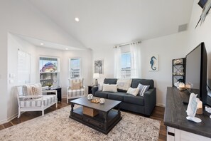 A living room with a dark wooden coffee table, a grey sofa, and a white armchair. There are windows with blinds, a bookshelf, and a television on a wooden stand.