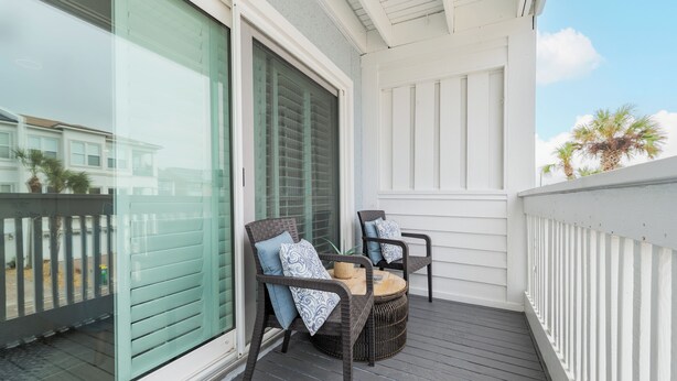Een balkon met twee rieten stoelen en een kleine ronde tafel, witte leuningen en een glazen deur.