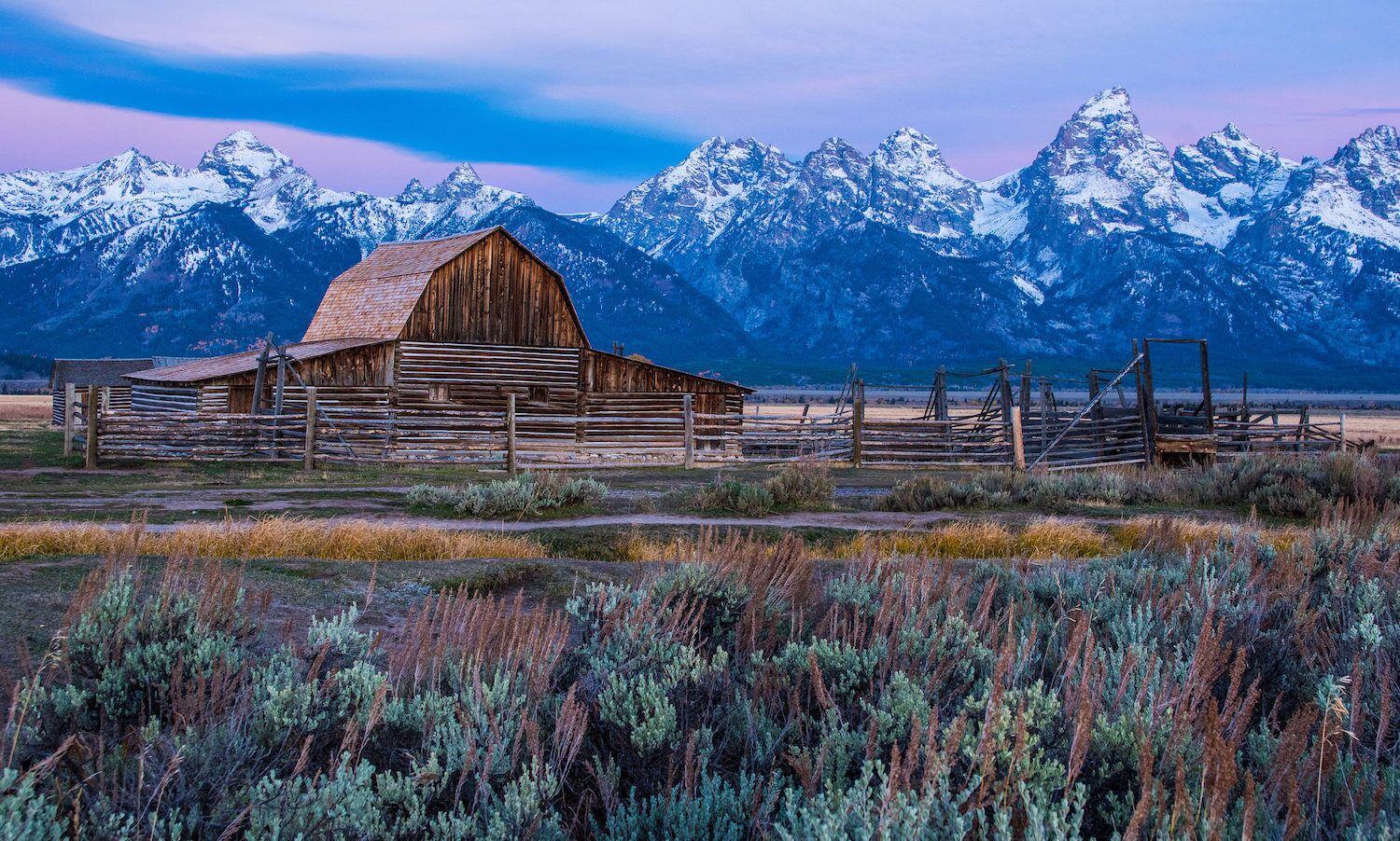 A rustic wooden barn with a sloped roof, surrounded by a wooden fence and a mountainous backdrop.