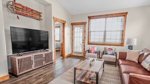 A living room with a large flat-screen TV, wooden entertainment unit, leather sofa, and a coffee table. - Teton National Park Avails! Great Location, Games, Laundry, & Playround! (Driggs)