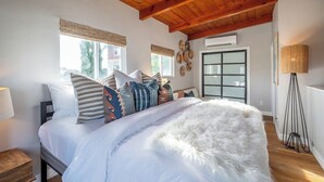 A bedroom with a wooden ceiling, a bed with white bedding and multiple pillows, a standing lamp, and a window with blinds.