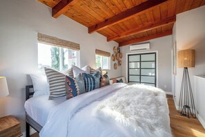 Une chambre avec un plafond en bois, un lit avec une literie blanche et plusieurs oreillers, une lampe sur pied et une fenêtre avec des stores.