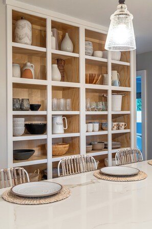 A dining area with a white table, multiple shelves with various kitchenware, and a hanging light fixture.