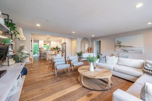 A spacious living area with a wooden floor, a round coffee table, and a sofa. There is a kitchen in the background with modern appliances and a dining area with chairs.