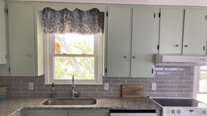 Cucina con mobili color verde chiaro, un piano di lavoro in granito, una finestra con tenda decorata, lavello in acciaio inossidabile, lavastoviglie bianca e fornello bianco.