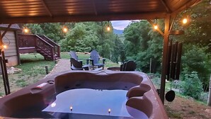 Un bain à remous installé sous une terrasse couverte, illuminée de guirlandes lumineuses, entouré d’une terrasse en bois et d’un chemin en pierre.