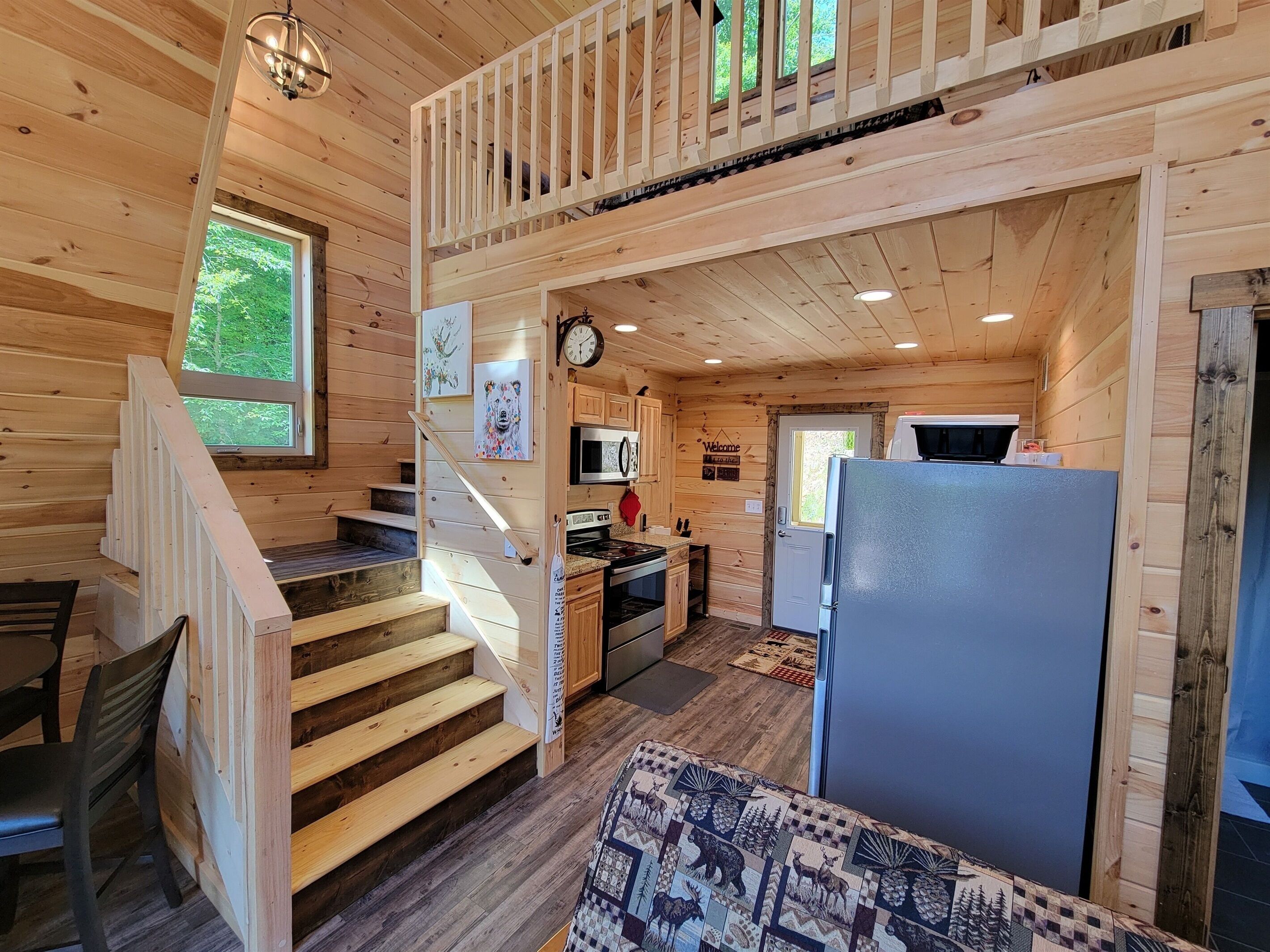 Interior de una cabaña de madera con una escalera que sube a un altillo, una zona de cocina con nevera y una sala de comedor con mesa y sillas.