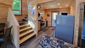 L’intérieur d’une cabane en bois comprend un escalier menant à une mezzanine, un coin cuisine avec un réfrigérateur et un coin repas avec une table et des chaises.