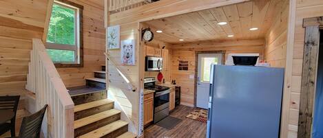 Interior de una cabaña de madera con una escalera que sube a un altillo, una zona de cocina con nevera y una sala de comedor con mesa y sillas.