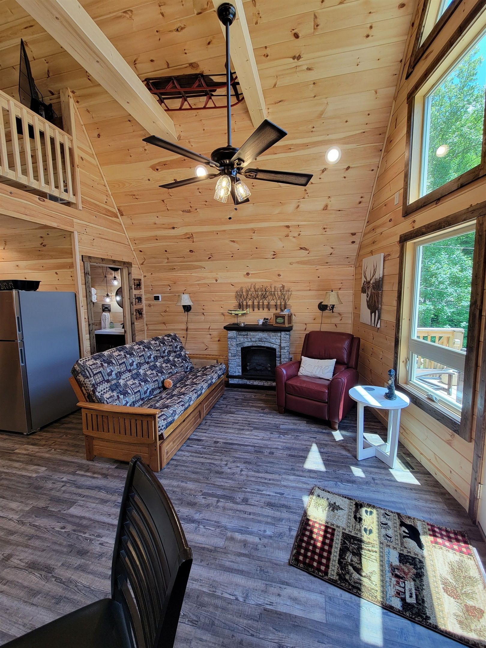 A cozy cabin interior with a wooden ceiling, a fireplace, a sofa, a red armchair, and a dining table.