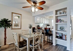 Un coin repas avec une table en verre, des chaises en osier, un ventilateur de plafond et une bibliothèque contenant des livres et des objets décoratifs.