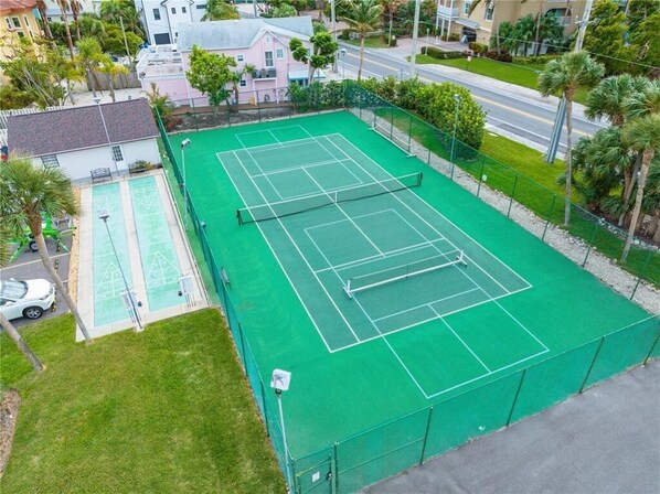 Una cancha de tenis con red, rodeada por una cerca verde, junto a una zona de piscina.