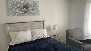Un lit bien fait, recouvert d’une couverture bleue, un fauteuil gris, une petite table de chevet avec une lampe, et un grand tableau floral accroché au mur.