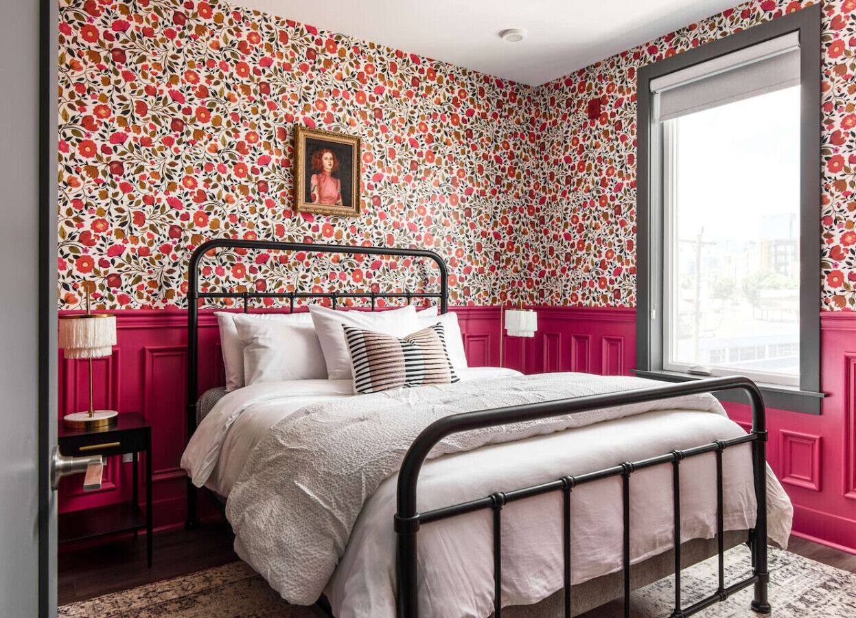A bedroom with a floral wallpaper, a bed with white bedding, a black metal frame, a bedside table with a lamp, and a window with a view of the outside.