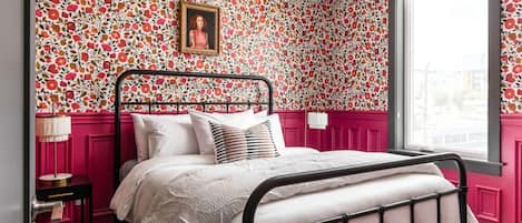 A bedroom with a floral wallpaper, a bed with white bedding, a black metal frame, a bedside table with a lamp, and a window with a view of the outside.