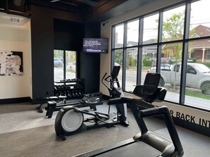 Un gimnasio con equipo cardiovascular, una televisión y grandes ventanales con vistas a la calle.