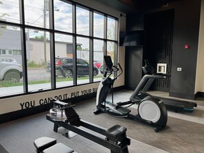 Un gimnasio con bicicletas estáticas, una cinta de correr y un amplio ventanal con vistas a un estacionamiento.