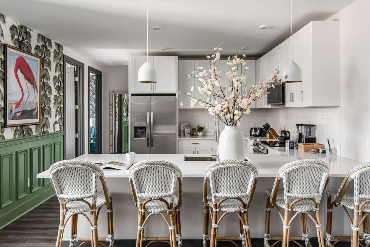 A modern kitchen with white cabinetry, a central island with bar stools, a large vase with flowers, and a refrigerator.