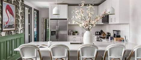A modern kitchen with white cabinetry, a central island with bar stools, a large vase with flowers, and a refrigerator.