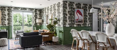 Una sala de estar moderna con una pared verde de acento, un ventanal grande y una zona de comedor con sillas de mimbre.
