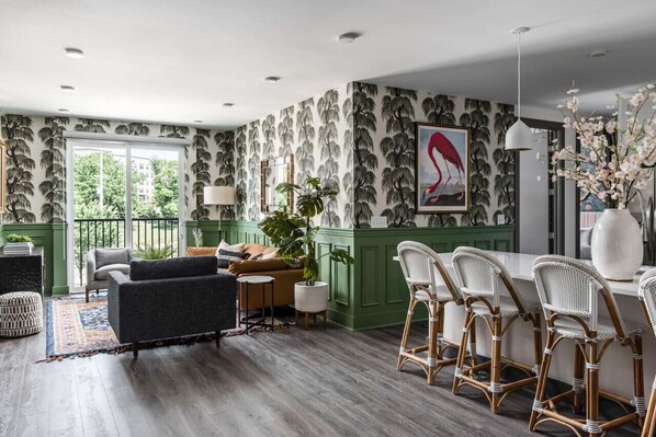 Una sala de estar moderna con una pared verde de acento, un ventanal grande y una zona de comedor con sillas de mimbre.