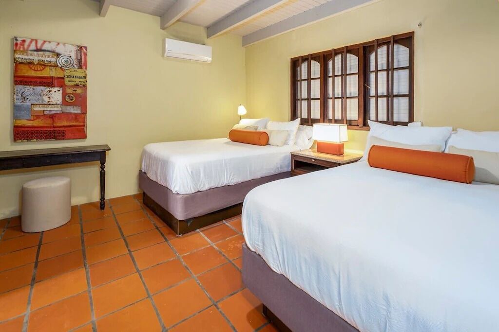 Een hotelkamer met twee bedden, een nachtkastje, een lamp, een raam met houten luiken en een luchtverwarmer aan de muur.