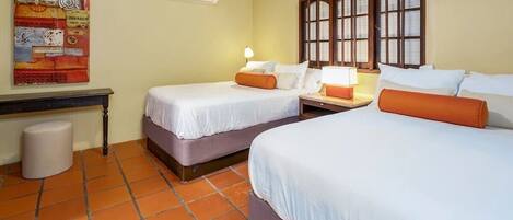 Een hotelkamer met twee bedden, een nachtkastje, een lamp, een raam met houten luiken en een luchtverwarmer aan de muur.