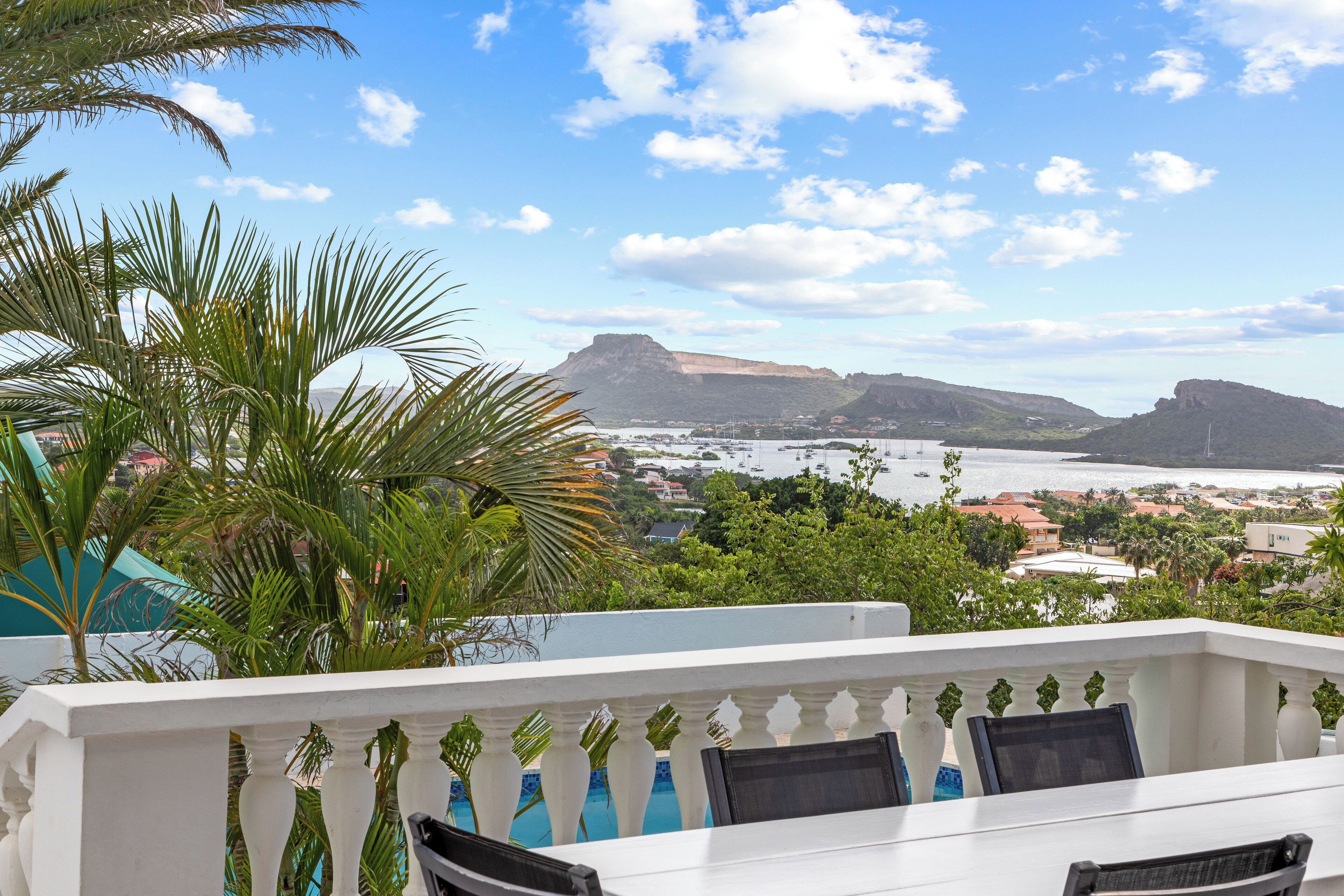 A balcony with white balustrades, a pool, and a view of a coastal town with mountains and a body of water.
