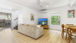 A modern living room with a beige sofa, a dining table with chairs, a flat-screen TV, and large windows with a view of greenery. - Peaceful Family Villa in Sunrise Beach (Sunrise Beach)