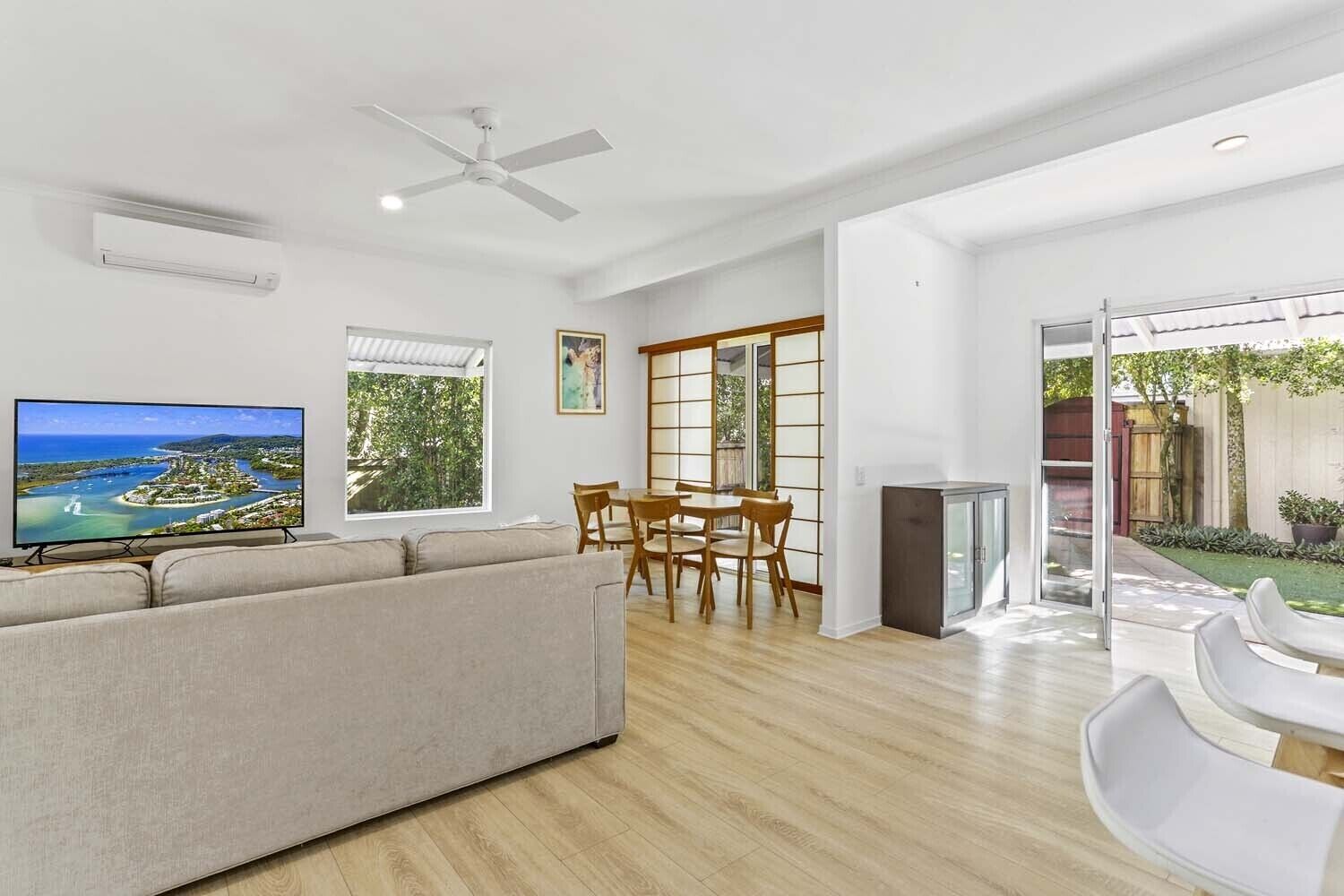 A spacious living area with a flat-screen TV, a ceiling fan, a dining table with chairs, and a view of a garden through large windows.