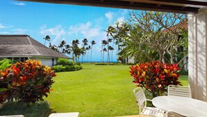 A covered outdoor area with a view of a lawn and palm trees, a building in the background, and a white table and chair set. - Luxury Ground Floor Condo Just 50 Yards From The Beach! (Koloa)
