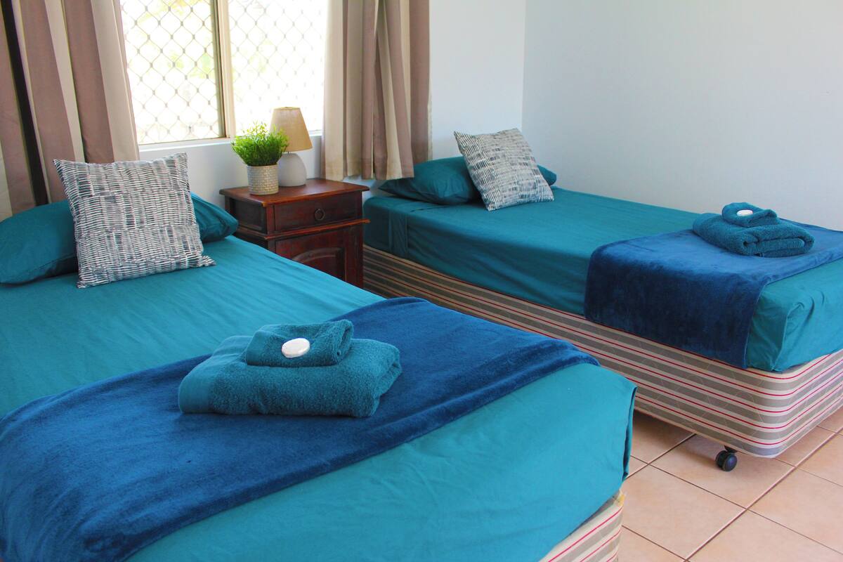 A hotel room with two beds, each with a teal bedspread and pillows, a small wooden nightstand with a lamp and a plant, and a window with curtains.