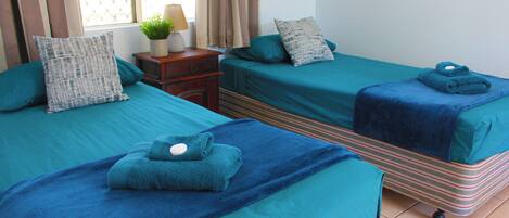 Habitación de hotel con dos camas, cada una con colcha y almohadas color turquesa, una mesita de noche de madera con lámpara y planta, y una ventana con cortinas.