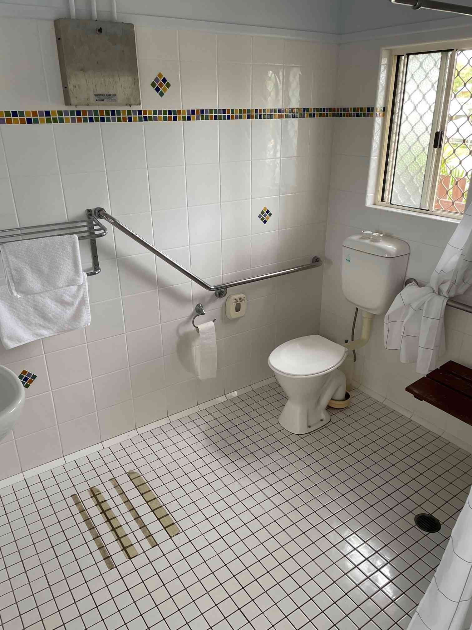 A bathroom with white tiled walls, a toilet, a handrail, a window, and a shower area.