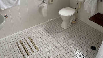 A bathroom with white tiled walls, a toilet, a handrail, a window, and a shower area.