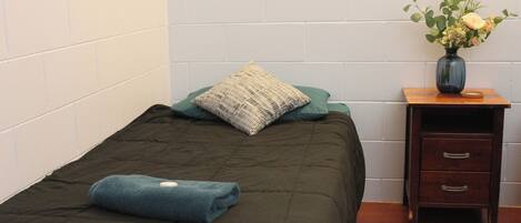A single bed with a dark green bedspread and a blue towel, a pillow, and a small wooden nightstand with a vase of flowers.