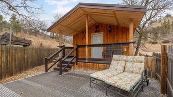 A small wooden cabin with a covered porch, a bed with a patterned mattress, and a staircase leading to the entrance.