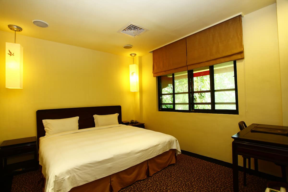 Ein Hotelzimmer mit Bett, Schreibtisch, Fenster mit Blick auf Grünflächen und Lüftungsgitter an der Decke.