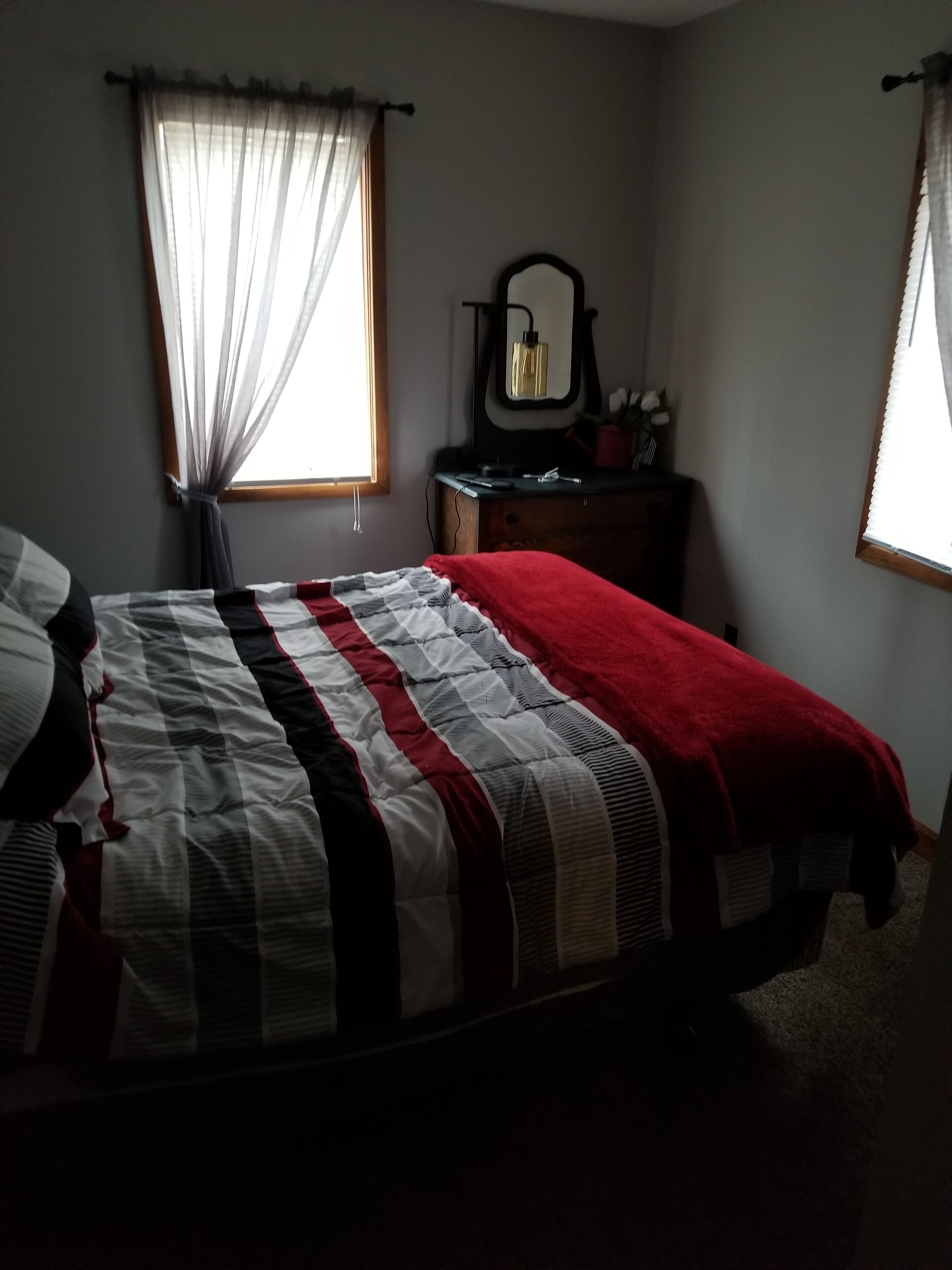 A bedroom with a bed, a dresser, a mirror, and a window with sheer curtains.