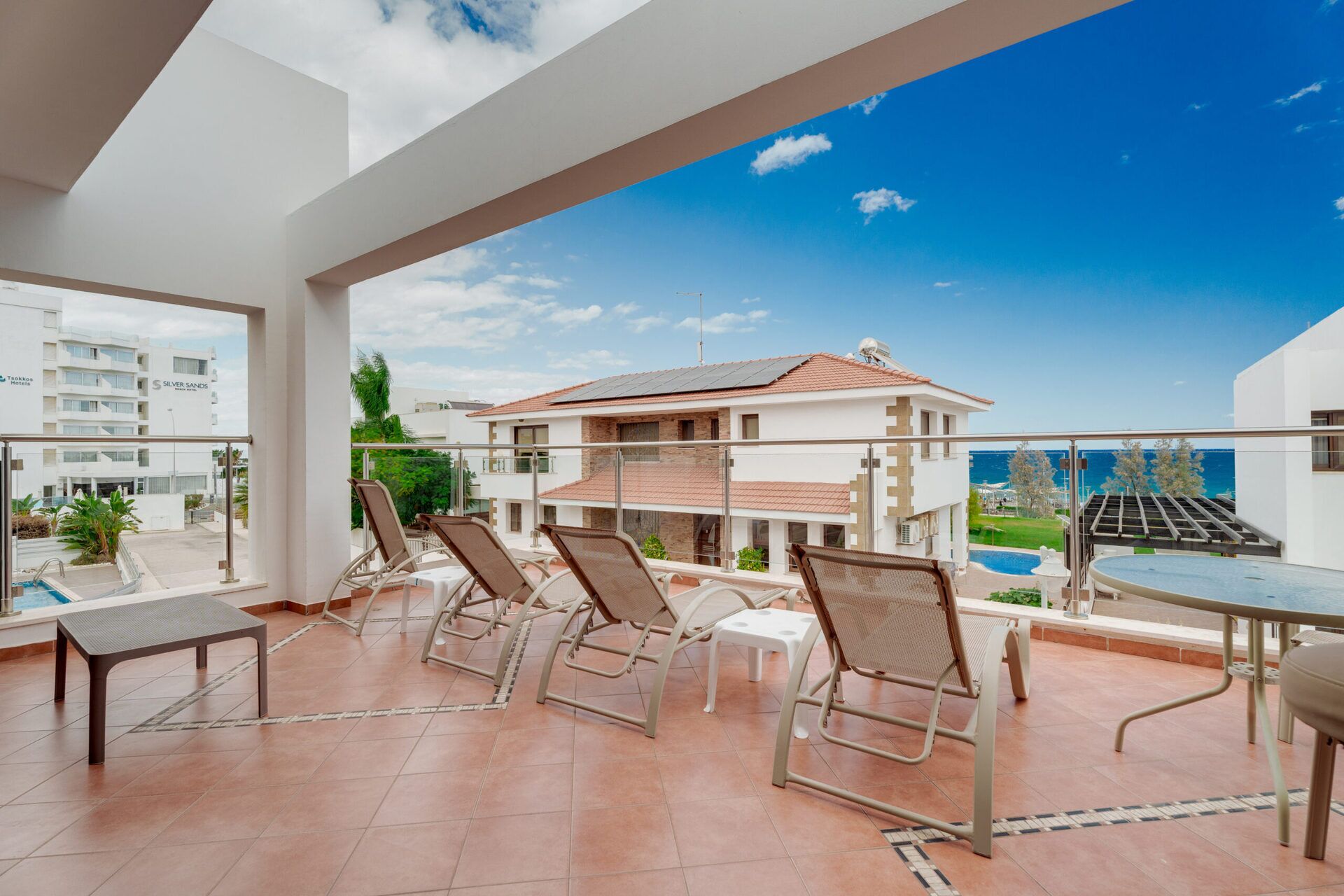 A balcony with lounge chairs and a pool, overlooking a building with a tiled roof and a view of the sea.
